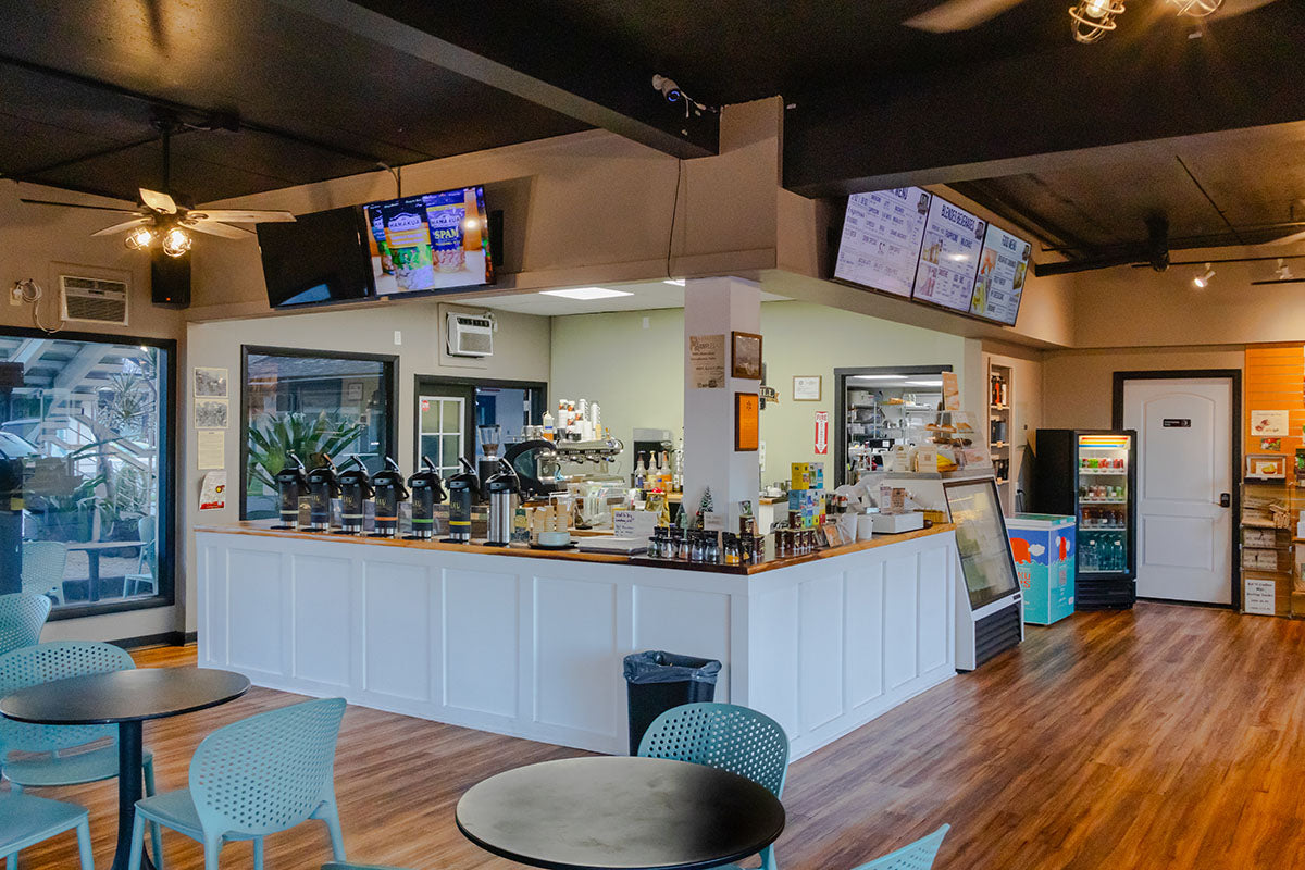 Kaʻū Coffee Roasters Cafe interior with a counter, seating area, and multiple screens on the wall.